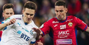 На изображении двое футболистов в форме команд: один в белой форме, другой в красной. Оба игрока сосредоточены на борьбе за мяч. На заднем плане видны размытые очертания стадиона и других игроков