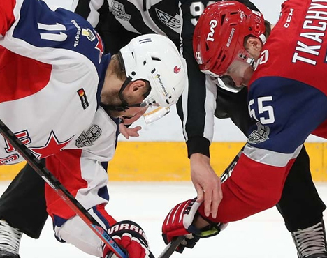 На изображении двое хоккеистов в разноцветной форме готовятся к вбрасыванию шайбы. Один в белой каске и форме, другой в красной. Между ними судья в черной форме. Ледовая арена видна на заднем плане