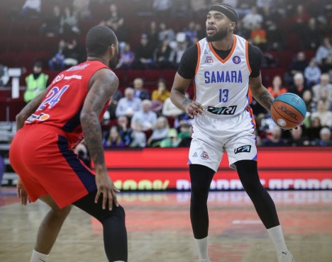 На изображении два баскетболиста на площадке. Игрок в белой форме с мячом готовится к игре против соперника в красной форме. На заднем плане видно размытую трибуну с сидящими зрителями